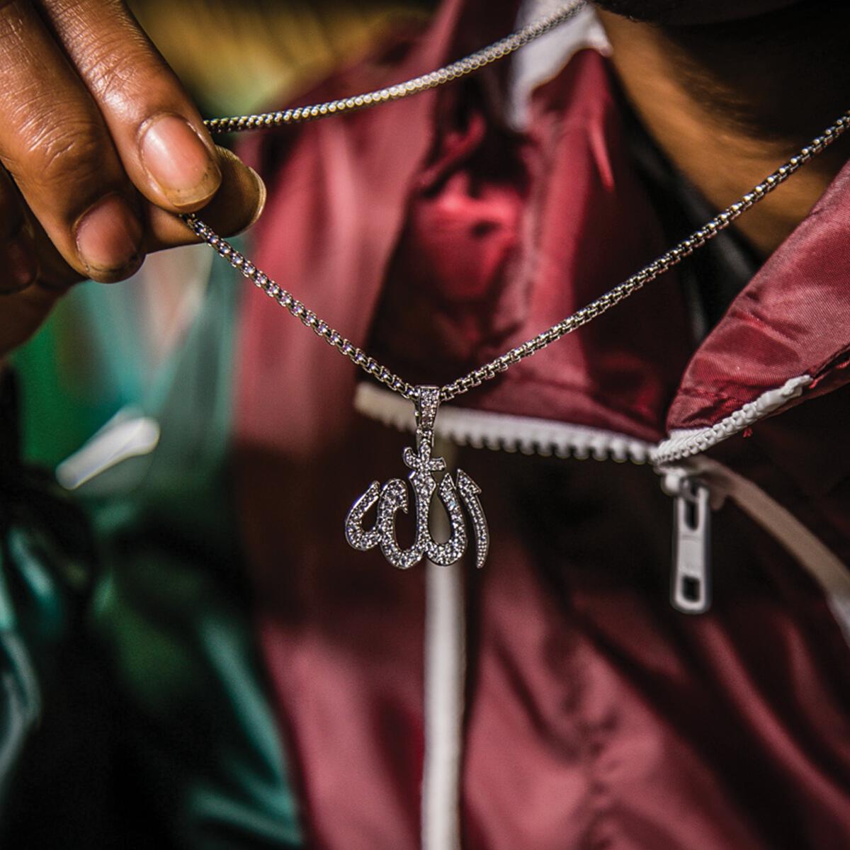Close-up of a silver chain with a pendant featuring Arabic calligraphy stating Allah, held by a hand.