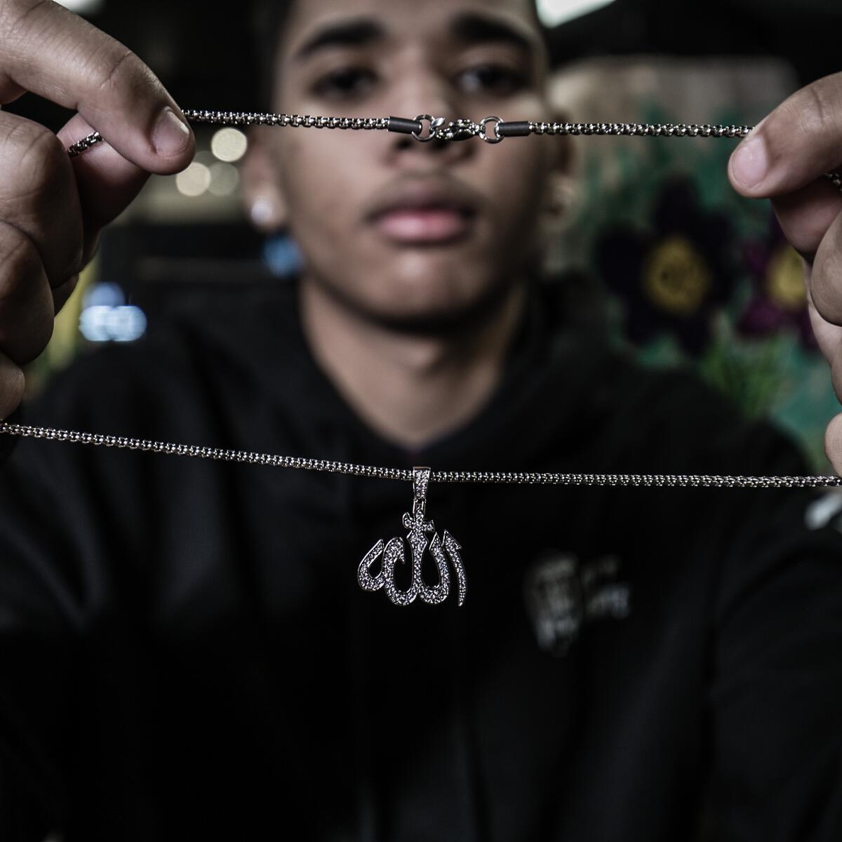 close up of a person in a Black hoodie holding a silver Allah necklace and text design on a dark background