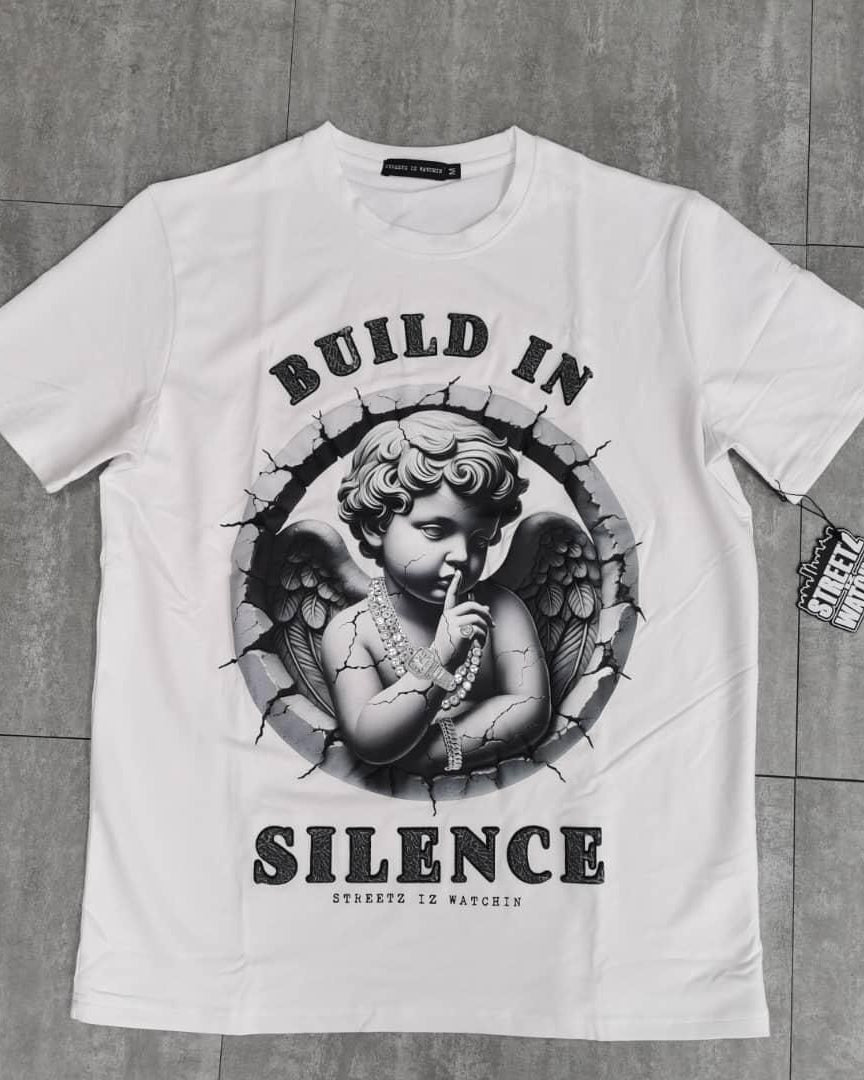 White t-shirt with an angel graphic and 'Build in Silence' text on a gray tiled floor.