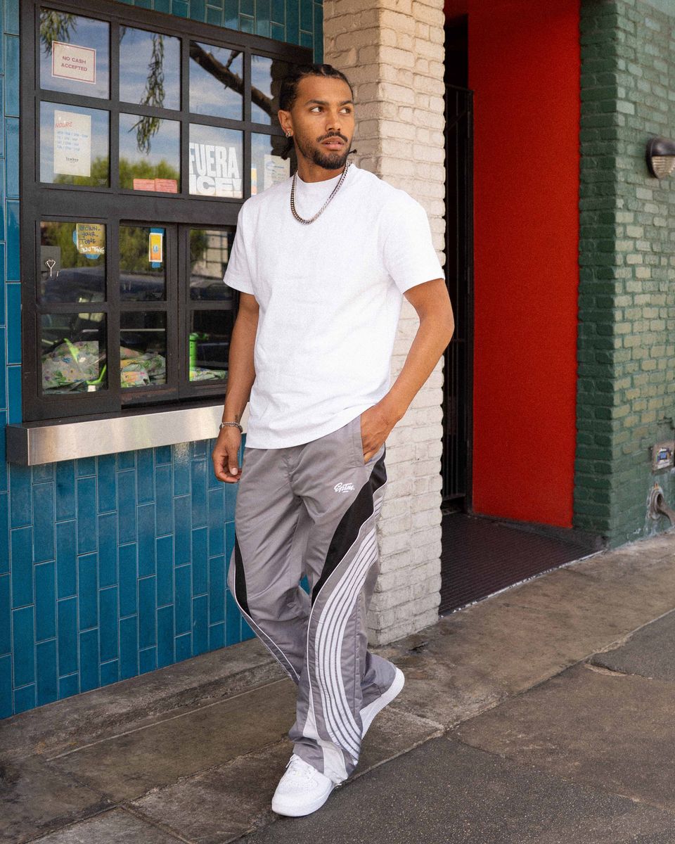 Man wearing a white t-shirt and gray pants standing outside a building with a blue tiled wall and red door.