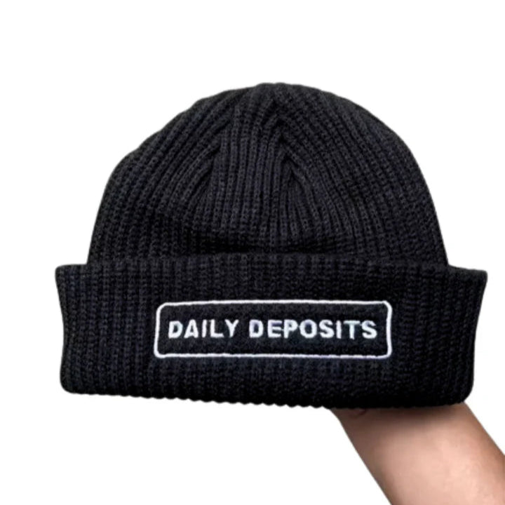 Black beanie with 'DAILY DEPOSITS' text held by a hand on a white background