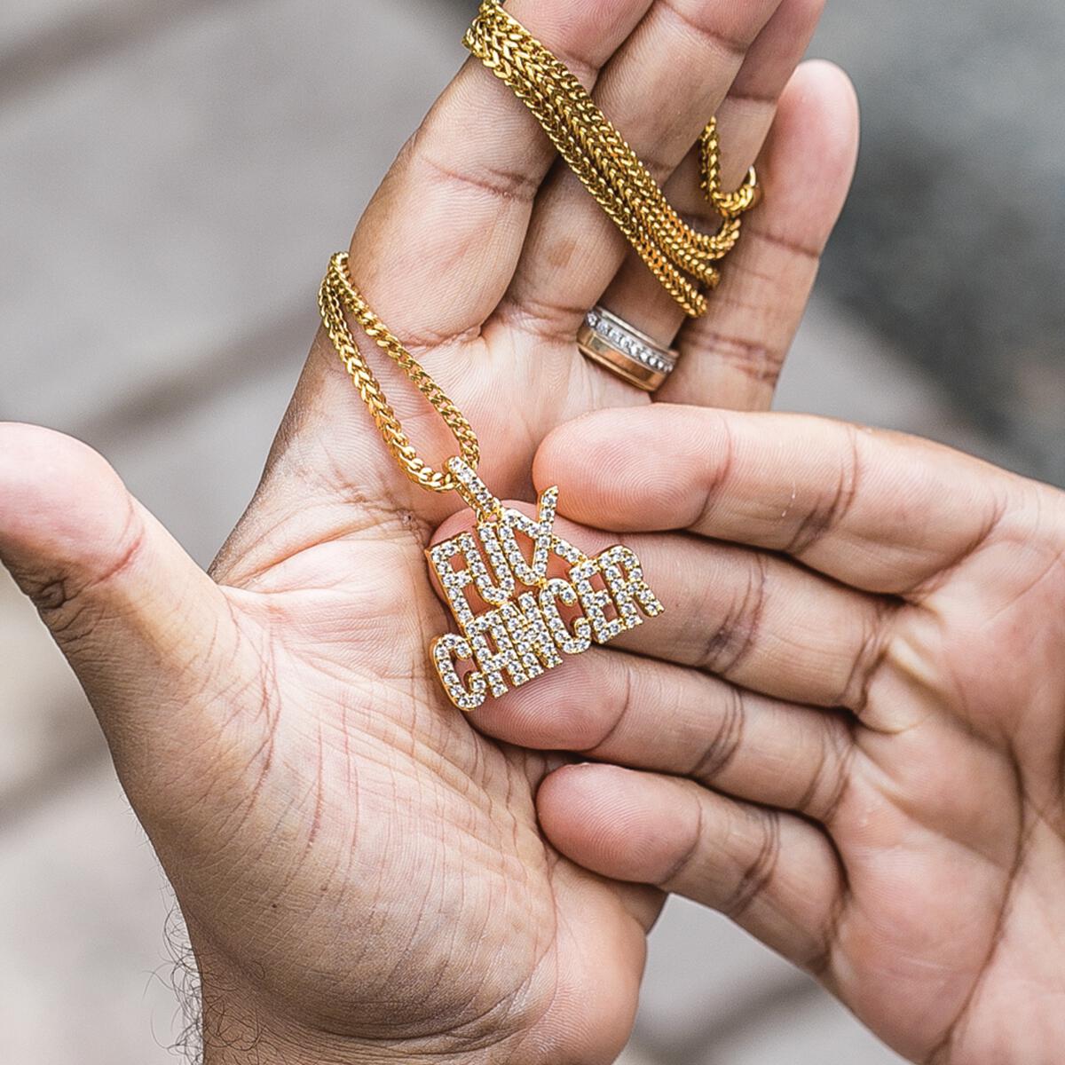Hand holding a gold chain with a 'Fucx Cancer' pendant against a neutral background