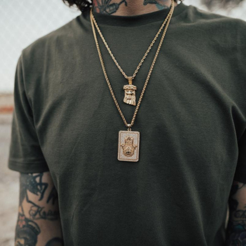 Person wearing a green t-shirt with gold necklaces featuring a mini Jesus and Hamsa hand pendants.