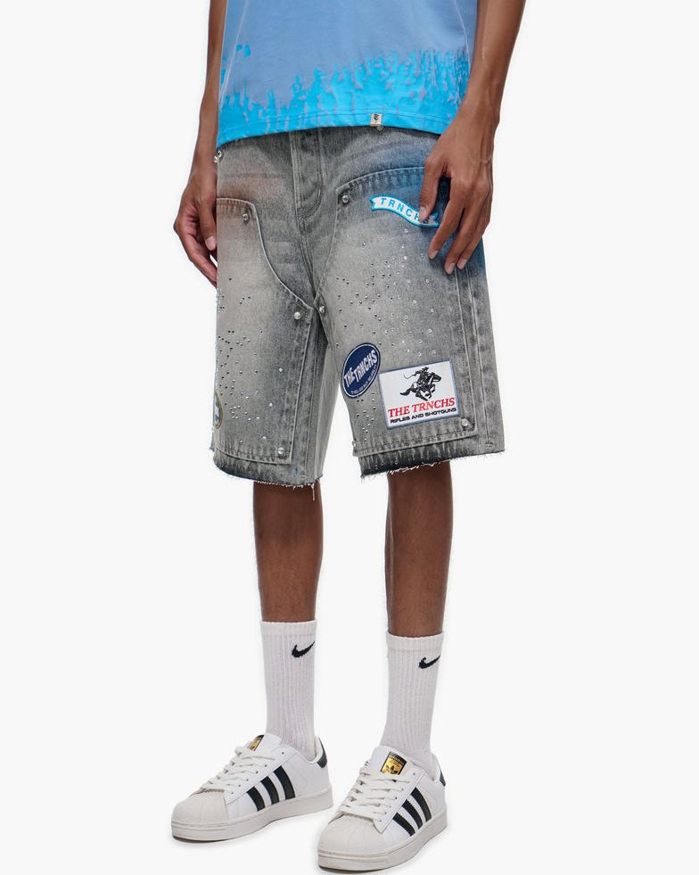 Person wearing gray denim shorts with patches, a blue top, white socks, and white sneakers on a white background.