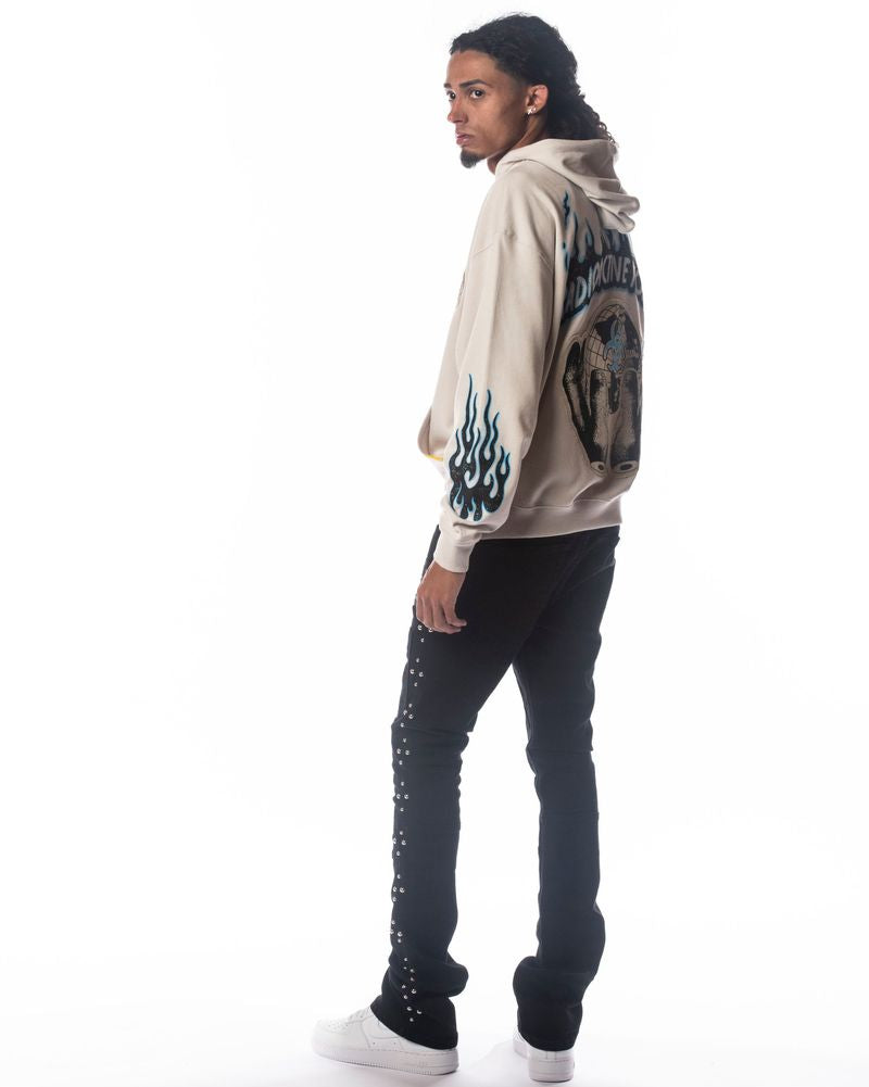 Person wearing a beige hoodie with graphic design on sleeves and black pants against a white background