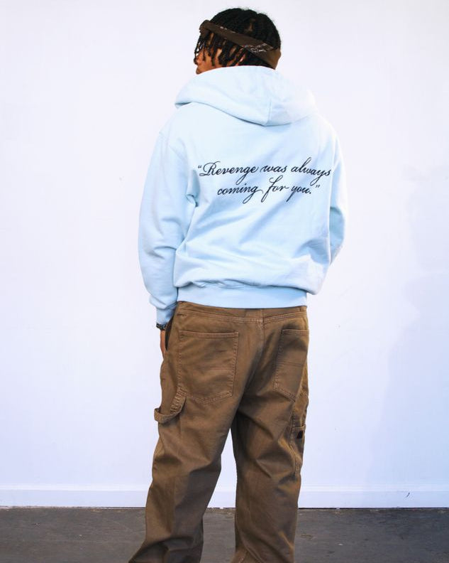 Person wearing a light blue hoodie with text on the back, standing against a white wall.