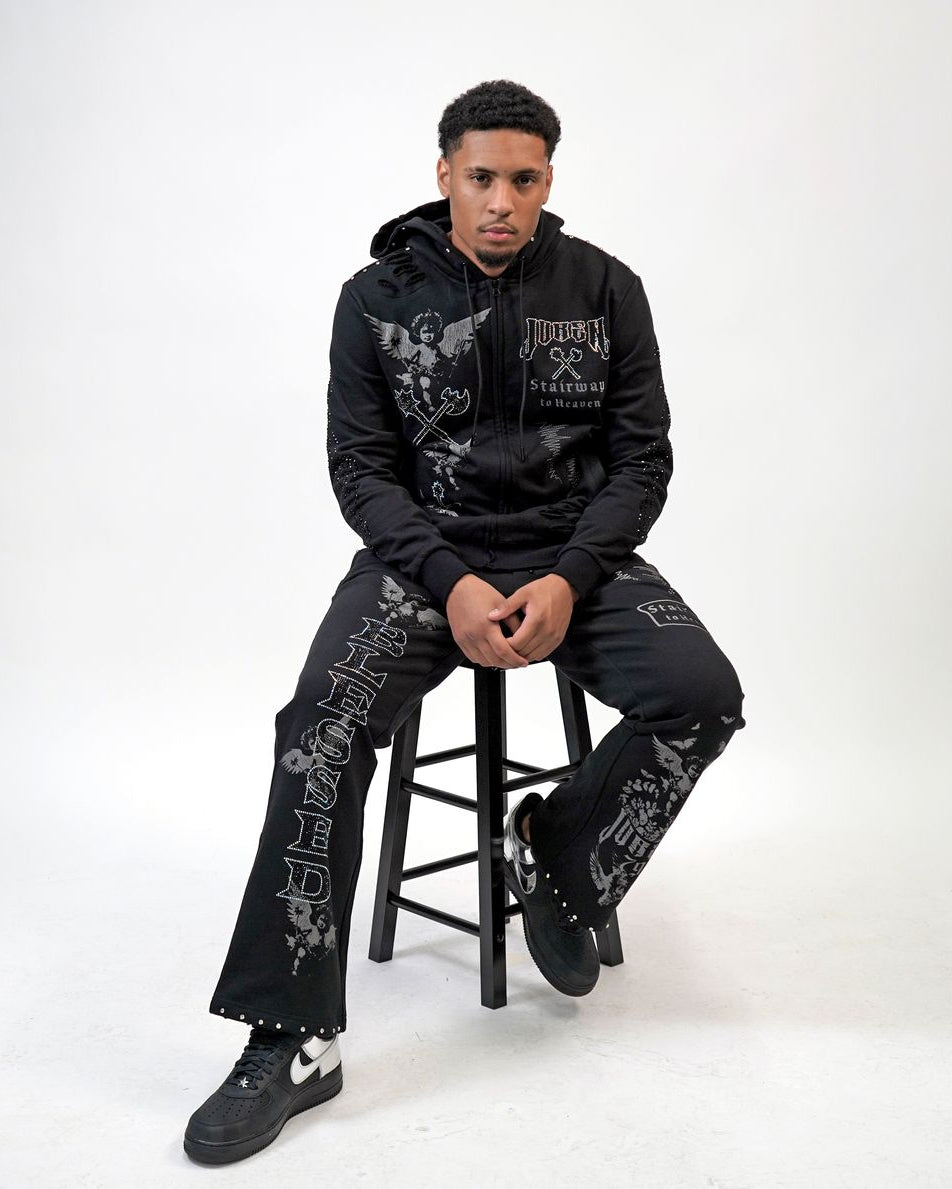 Person wearing a black hoodie and pants with graphic designs, sitting on a stool against a white background.