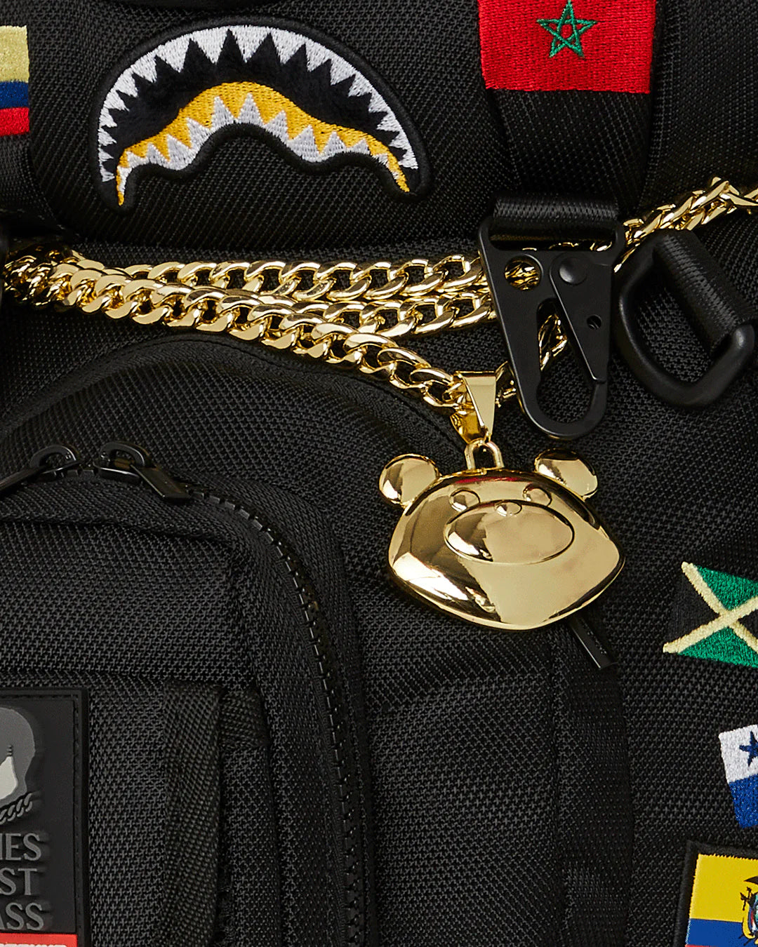Black backpack with gold chain and bear pendant, featuring various patches including a shark logo and flags.
