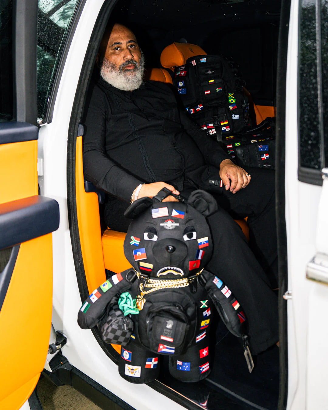 Man sitting in a vehicle with a decorative black bear plush toy featuring international flags.