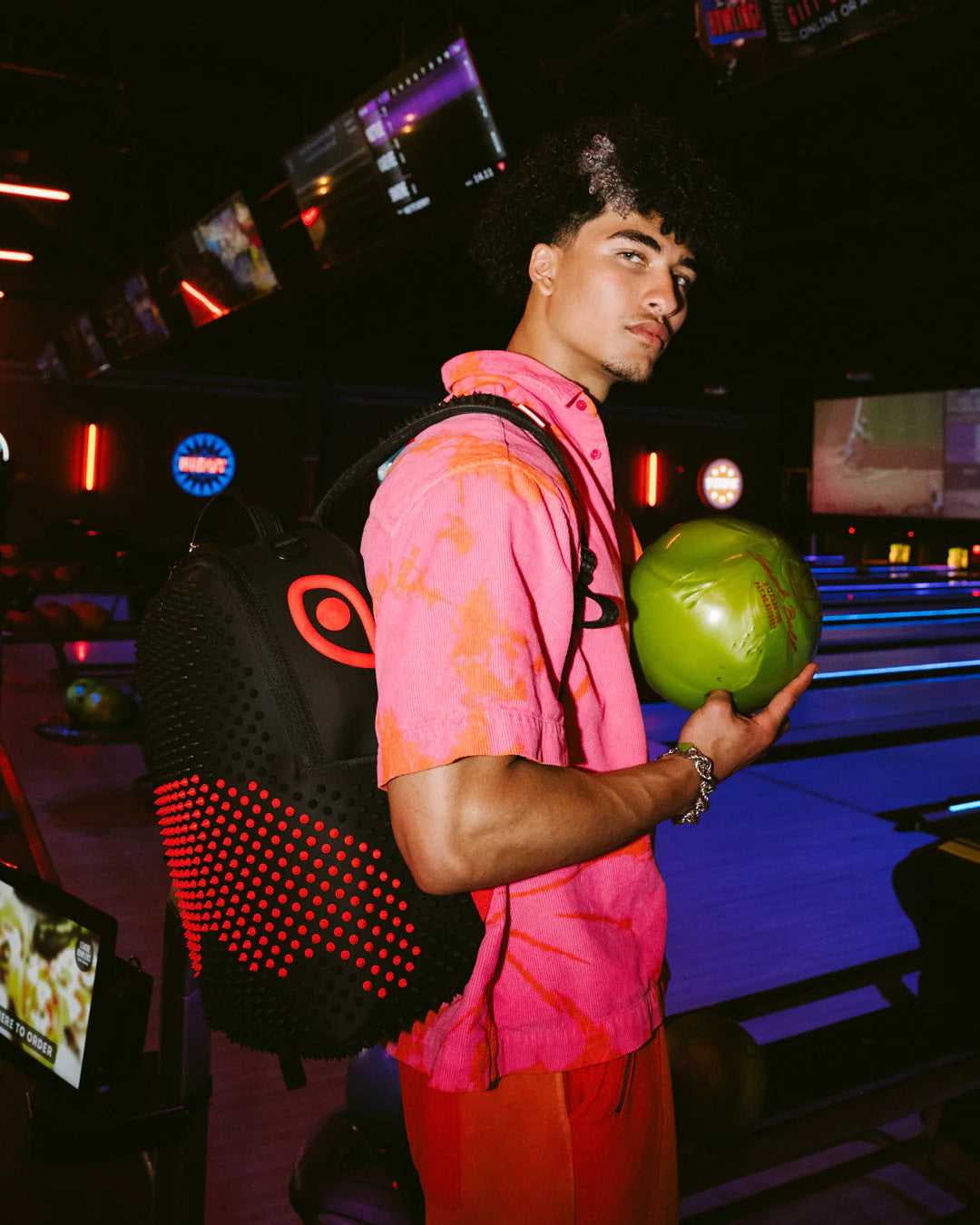 Person in a pink shirt holding a green ball in a bowling alley wearing the black backpack with red and black spikes by sprayground