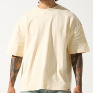 A male model wearing a beige crew neck t-shirt with short sleeves and distressed detailing at the neck and sleeves. The t-shirt has a crew neckline and a slightly cropped fit.