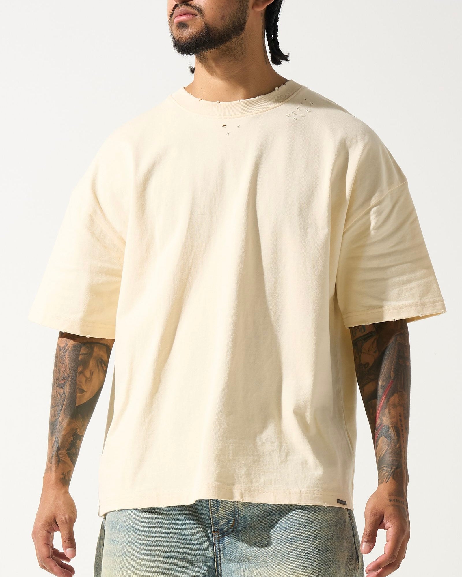 A male model wearing a beige crew neck t-shirt with short sleeves and distressed detailing at the neck and sleeves. The t-shirt has a crew neckline and a slightly cropped fit.