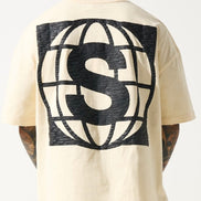 A person wearing a beige crew neck t-shirt with a large 'S' logo on the back, and distressed detailing on the neck and sleeves.