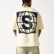 A male model wearing a beige crew neck t-shirt with short sleeves and distressed detailing at the neck and sleeves. The t-shirt has a crew neckline and a slightly cropped fit.