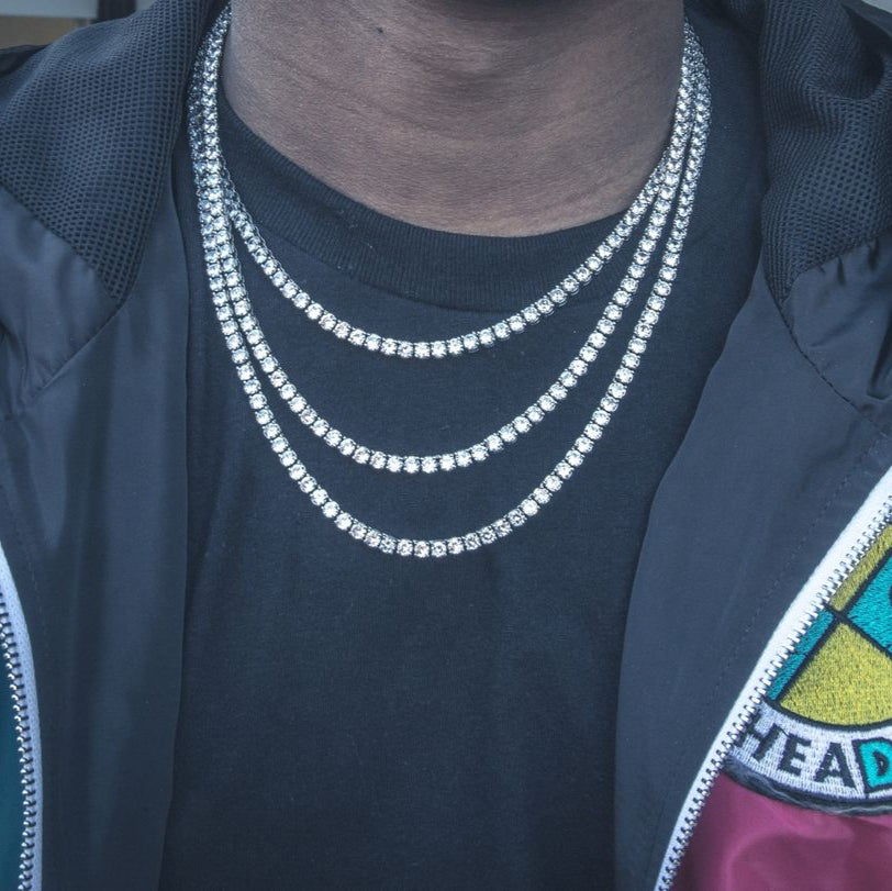 Person wearing a dark jacket with a colorful patch and multiple tennis necklaces.