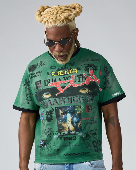 Man wearing a green graphic t-shirt with various designs and text on a plain background