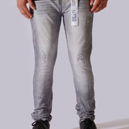 A person wearing light gray washed denim jeans with distressed details, paired with a black t-shirt and black sneakers, shown from the waist down against a light gray studio background.