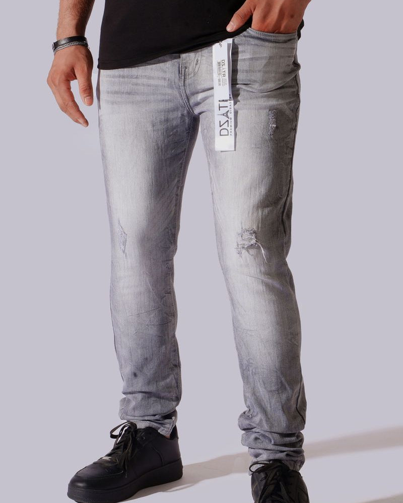 A person wearing light gray washed denim jeans with distressed details, paired with a black t-shirt and black sneakers, shown from the waist down against a solid gray background.