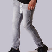 A person wearing light gray washed denim jeans with a distressed knee, paired with a black t-shirt and black sneakers, shown from the waist down against a solid light gray background.