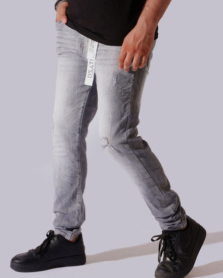 A person wearing light gray washed denim jeans with a distressed knee, paired with a black t-shirt and black sneakers, shown from the waist down against a solid light gray background.