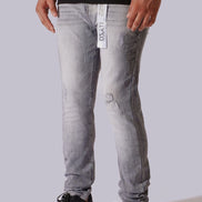 A person wearing light gray washed skinny jeans with a visible hangtag, paired with a black shirt and black sneakers, against a plain light gray studio background.