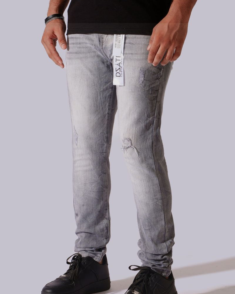 A person wearing light gray washed skinny jeans with a visible hangtag, paired with a black shirt and black sneakers, against a plain light gray studio background.