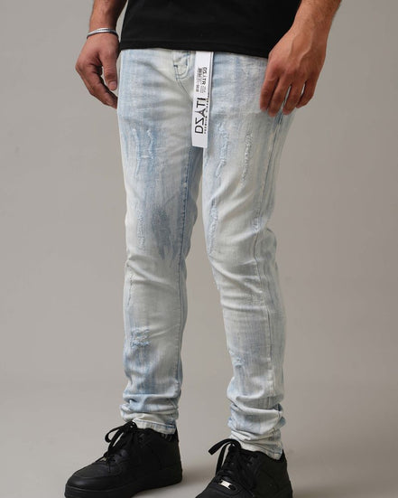Person wearing light blue distressed jeans with a brand label and black shoes on a gray background