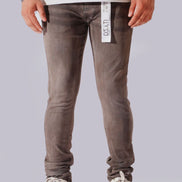 A person from the waist down wearing slim-fit gray wash jeans with white sneakers, shown against a plain light gray studio background. A white hangtag is visible on the right side of the jeans.