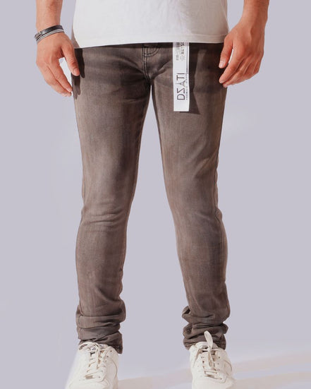A person from the waist down wearing slim-fit gray wash jeans with white sneakers, shown against a plain light gray studio background. A white hangtag is visible on the right side of the jeans.