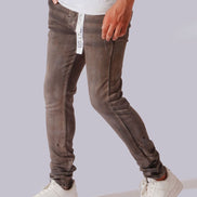 A person wearing slim-fit brown washed jeans with a white t-shirt and white sneakers, shown from the waist down against a light gray background.