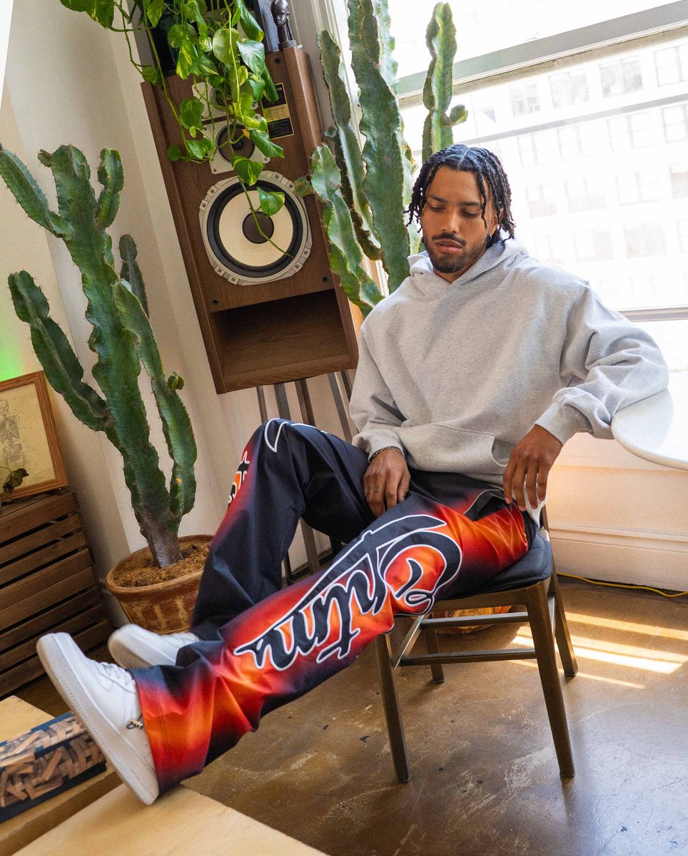 Person sitting on a chair wearing colorful pants with a brand logo, surrounded by plants and a speaker.