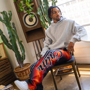 Person sitting in a room with plants and a speaker, wearing colorful pants and a gray hoodie.