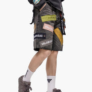 Person wearing a black jacket with patches and gray shorts on a white background