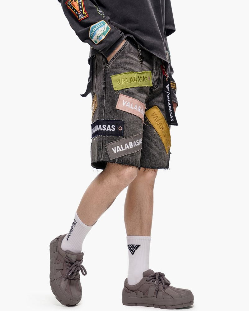 Person wearing a black jacket with patches and gray shorts on a white background