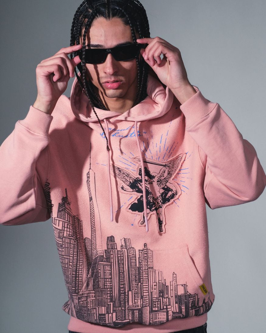 Person wearing a pink hoodie with cityscape design and sunglasses, against a gray background