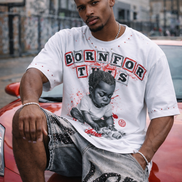 Man wearing a graphic t-shirt and shorts, sitting on a red car in an urban setting.