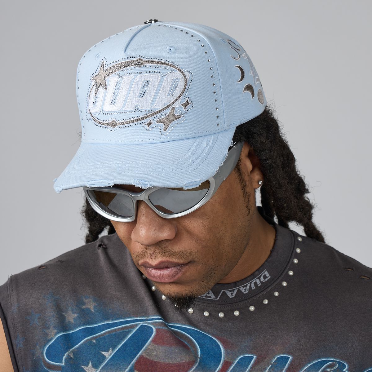 A man wearing a light blue baseball cap with a silver and white 'DUAA' logo, silver sunglasses, and a dark gray graphic t-shirt against a solid gray background.