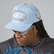 A man wearing a light blue distressed baseball cap with silver rhinestone 'DUAA' lettering on the front and silver sunglasses, shown in a close-up profile shot against a neutral gray background.