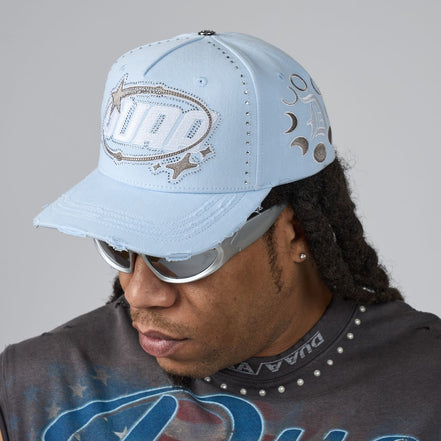 A man wearing a light blue distressed baseball cap with silver rhinestone 'DUAA' lettering on the front and silver sunglasses, shown in a close-up profile shot against a neutral gray background.