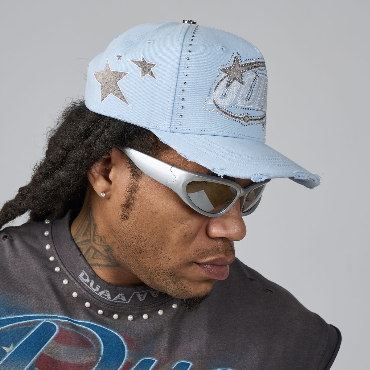 A man wearing a light blue distressed baseball cap with rhinestone star and logo details, white sunglasses, and a dark gray graphic t-shirt against a solid gray background.