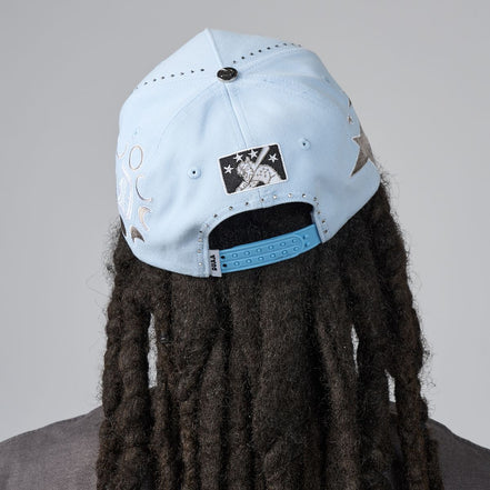 A light blue baseball cap with silver graphic prints and a black-and-white patch, shown from the back on a person with long dark dreadlocks against a gray background.
