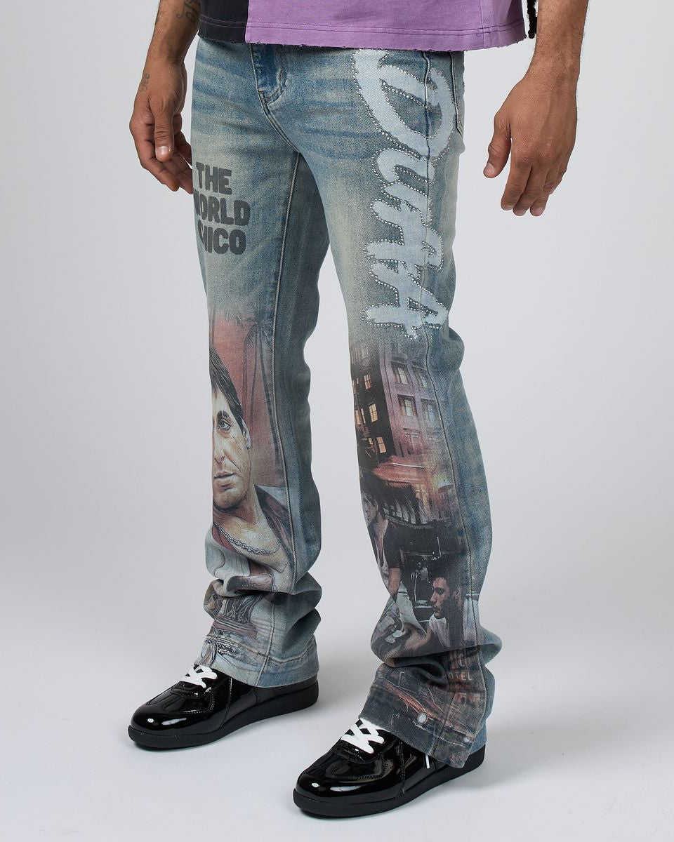 A person wearing blue denim jeans with large graphic prints and text, paired with black lace-up shoes, photographed against a light gray studio background.