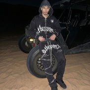 Person wearing a black hoodie with white text standing next to a vehicle in a desert setting at night.