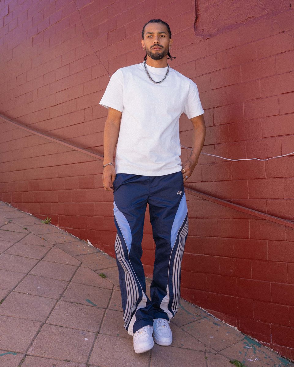 Man wearing a white t-shirt and blue tracksuit standing against a red brick wall.