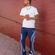 Man wearing a white t-shirt and blue tracksuit with white stripes against a red brick wall.