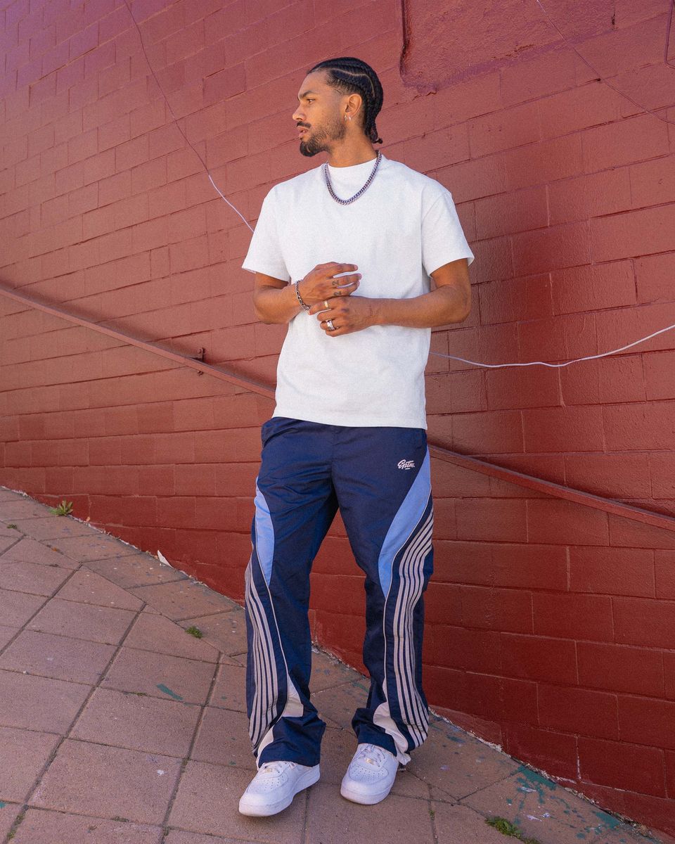 Man wearing a white t-shirt and blue tracksuit with white stripes against a red brick wall.