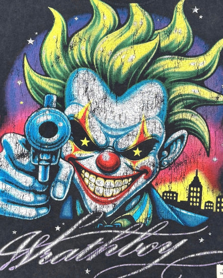 A close-up graphic print featuring a stylized clown with bright green hair, a red nose, and a blue suit holding a revolver, with the word 'Whathavey' in white script across the bottom.