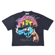 A dark black graphic t-shirt featuring a cartoon-style blue revolver with a surprised face, a brown gun handle, and a pink and yellow city skyline under the word 'Wanted' in white script.