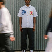 Person wearing a white shirt with logos standing against a corrugated metal wall.