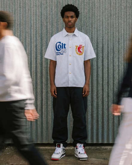 Person wearing a white shirt with logos standing against a corrugated metal wall.